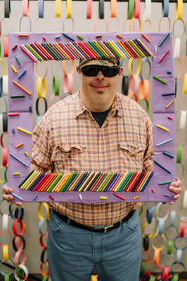 Guy holding a frame for a picture with colored pencils on it